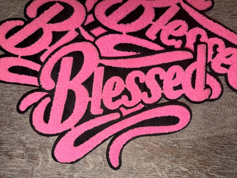 Puede incluir: Primer plano de un parche rosa y negro con la palabra "Blessed" en cursiva. El parche tiene una textura difusa y est&aacute; sobre un fondo de madera. La palabra "Blessed" es el foco principal.