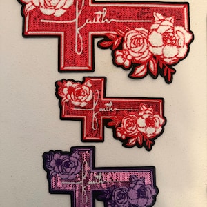 May include: Three cross-shaped patches with floral designs and the word "faith". The top two are red with white flowers, and the bottom one is purple with purple flowers. Each patch has a black border.