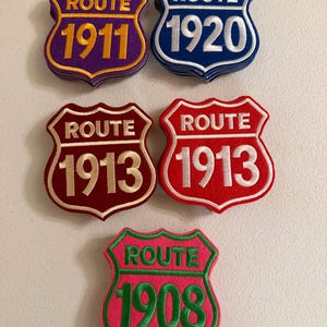 May include: Five stacks of shield-shaped embroidered patches featuring the word ROUTE and various years. The patches include purple with 1911, blue with 1920, dark red with 1913, bright red with 1913, and a pink patch with green text reading ROUTE 1908.