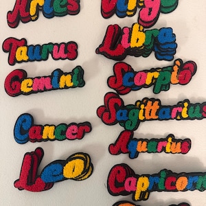 May include: A collection of twelve embroidered patches featuring the names of the zodiac signs: Aries, Taurus, Gemini, Cancer, Leo, Virgo, Libra, Scorpio, Sagittarius, Aquarius, Capricorn, and Pisces, each in multi-colored, puffy lettering.