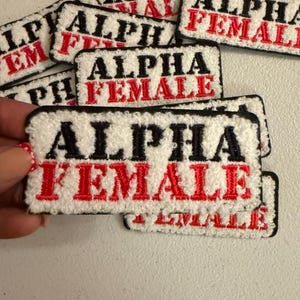 May include: A collection of rectangular embroidered patches with a white textured background. Each patch features the word ALPHA in black capital letters on the top line and the word FEMALE in red capital letters on the bottom line, with black borders.