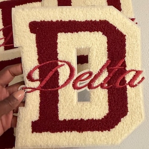 May include: A large, cream-colored letter D with a textured, fuzzy surface and a dark red border. The word Delta is embroidered in dark red cursive script across the center of the letter. The item features a thick, plush pile construction.