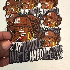May include: A collection of embroidered patches featuring a person wearing a brown baseball cap with the letters ATL, gold hoop earrings, and long white nails covering their mouth. The text STAY HUMBLE HUSTLE HARD is embroidered in white and gray.