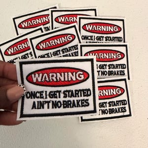 May include: A collection of rectangular embroidered patches featuring a white background with a black border. Each patch displays a red oval containing the white text WARNING, followed by the black text ONCE I GET STARTED AIN'T NO BRAKES.