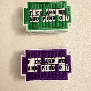 May include: Two rectangular embroidered patches featuring a grid design. One patch is green and the other is purple. Both patches contain white squares with black letters spelling out the phrase FUCK AROUND AND FIND OUT in a crossword style.