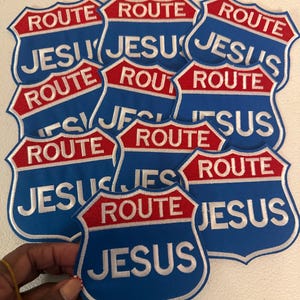 May include: A collection of shield-shaped embroidered patches featuring a blue background with a red top banner. The word ROUTE is stitched in white on the red banner, and the word JESUS is stitched in white on the blue section below.