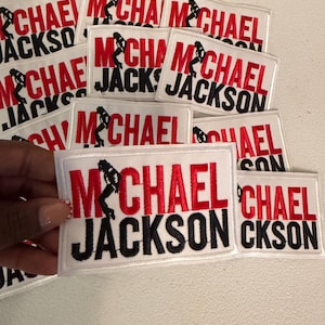 May include: A collection of rectangular white embroidered patches featuring the text Michael Jackson in red and black thread. Each patch includes a small black silhouette of a dancer in a signature pose integrated into the letter M.