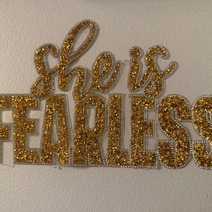 May include: A gold glitter applique with the words "she is FEARLESS". The letters are outlined in silver and are on a white background. The applique is likely for clothing or crafts.