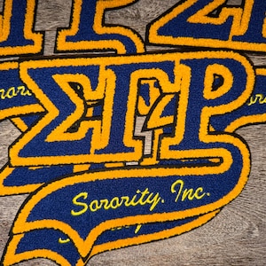 May include: Close-up of several embroidered patches in blue and gold. The patches feature Greek letters and the words "Sorority, Inc." in yellow script. The patches are likely for a sorority or similar organization.