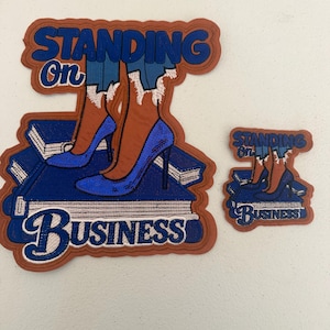 May include: Two patches with the text "STANDING ON BUSINESS". The larger patch features a pair of blue high heels on a stack of books. The smaller patch has the same design. Both patches have a brown border.