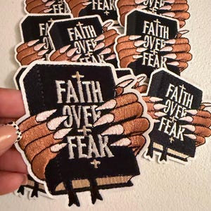May include: A collection of embroidered patches featuring a black book with the white text FAITH OVER FEAR. The design depicts a pair of tan-colored hands with long, light-colored fingernails holding the book, which includes small cross symbols.