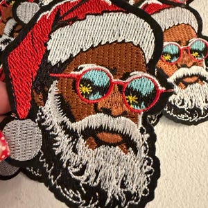 May include: An embroidered patch featuring a Santa Claus figure with brown skin, a white beard, and a red hat. The figure wears round, red-rimmed sunglasses with light blue lenses that have small yellow star-like reflections on the surface.