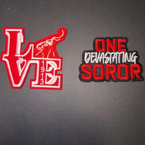 May include: Two embroidered patches. The first patch spells "LOVE" in red with white trim, featuring an elephant graphic and the numbers "1913". The second patch reads "ONE DEVASTATING SOROR" in red and white with a black border.