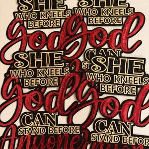 May include: Multiple fabric patches featuring the text 'SHE WHO KNEELS BEFORE GOD CAN STAND BEFORE ANYONE'. The words 'SHE WHO KNEELS BEFORE' and 'CAN STAND BEFORE' are embroidered in gold thread on black fabric, while 'GOD' and 'ANYONE' are in red sequins.
