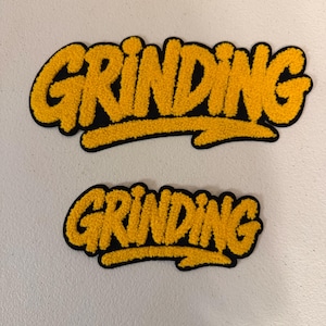 May include: Two embroidered patches featuring the word GRINDING in a bold, yellow, textured font with a black outline. The patches are identical in design, with one significantly larger than the other, showcasing a graffiti-inspired lettering style.