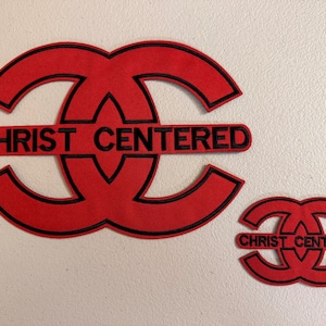 May include: Two red and black embroidered patches featuring a stylized interlocking circular design. The larger patch on the left and the smaller patch on the right both display the text CHRIST CENTERED in black capital letters across the center.