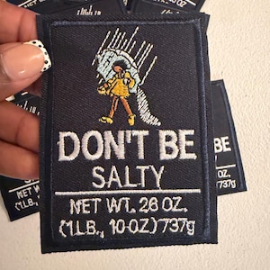 May include: A rectangular black embroidered patch featuring a graphic of a person in a yellow coat holding an umbrella with salt pouring out. The text reads DON'T BE SALTY, NET WT. 26 OZ. (1LB., 10 OZ.) 737g in white thread on a dark background.