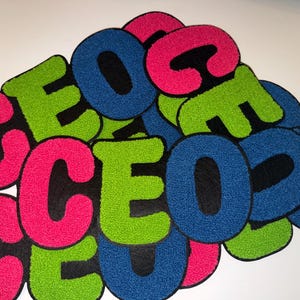 May include: A collection of colorful, plush letter patches. The letters are in a bold, block style with a black backing. The letters are in bright pink, green, and blue. The letters are arranged in a pile.