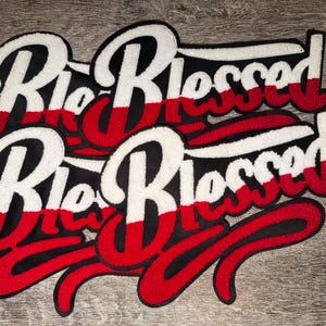 May include: Embroidered patches with the text "Rio Blessed" in a stylized script. The patches feature a color scheme of white, red, and black, with a textured, raised appearance. The patches are likely for clothing or accessories.