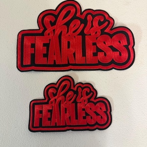 May include: Two red embroidered patches with the words "she is FEARLESS" in a cursive font. The text is outlined in black, with a black border. One patch is larger than the other.