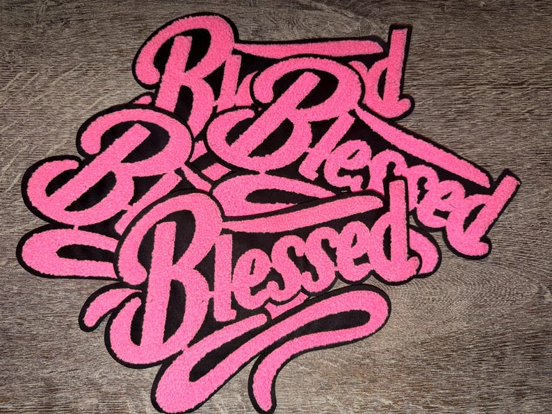Puede incluir: Un parche bordado en rosa y negro con la palabra "Blessed" en cursiva. El parche tiene un fondo negro y un contorno rosa y difuso. La palabra "Blessed" se repite varias veces.