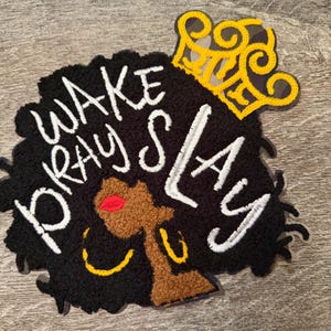 May include: Black and brown decorative patch featuring a stylized woman's face with the text "WAKE PRAY SLAY" in white. The woman is wearing gold hoop earrings and a gold crown. The patch has a textured appearance.