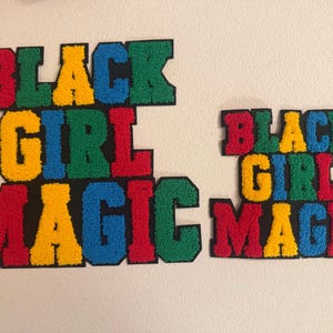 May include: Two colorful embroidered patches with the words "BLACK GIRL MAGIC". The patches feature a black background with letters in red, green, yellow, and blue. One patch is larger than the other.