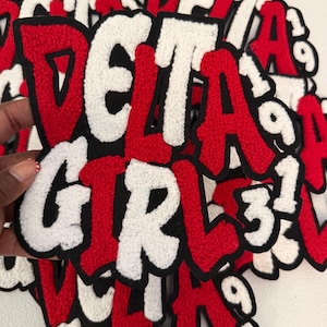 May include: A collection of embroidered patches featuring the text DELTA GIRL in a bold, layered font. The letters alternate between red and white chenille textures, each outlined in black. The patches are stacked, showing multiple identical designs.