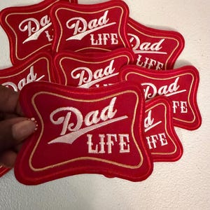 May include: A collection of red embroidered patches with a scalloped rectangular shape. Each patch features a gold border and the white stitched text "Dad LIFE" in a stylized font. The design mimics a classic beverage label aesthetic.