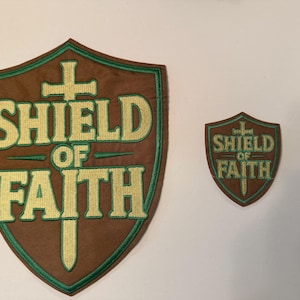 May include: Two brown shield-shaped patches with the text "SHIELD OF FAITH" in gold and green embroidery. The larger patch is approximately 6 inches tall, and the smaller one is about 3 inches tall. The patches have a green border.