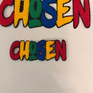 May include: Two tufted wall hangings featuring the word CHOSEN in a bold, rounded font. Each letter is a different color, including red, yellow, green, and blue, with a thick black outline surrounding the entire word and individual letters.