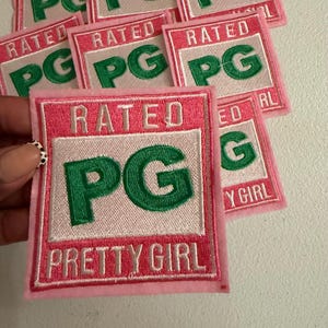 May include: A rectangular embroidered patch featuring a pink border and a white center. The text RATED is embroidered in white at the top, the letters PG are in large green font in the center, and the words PRETTY GIRL are in white at the bottom.