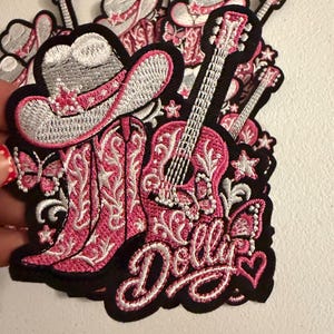 May include: A stack of embroidered patches featuring a silver cowboy hat, pink cowboy boots, and a pink acoustic guitar. The design includes butterflies, stars, and a heart, with the word Dolly written in white cursive script on a black background.