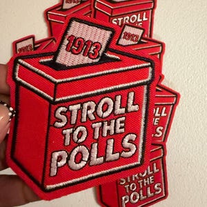 May include: A red embroidered patch shaped like a ballot box with a white ballot inserted at the top. The ballot features the red text 1913. The box displays the white text STROLL TO THE POLLS. The design is outlined in black thread.