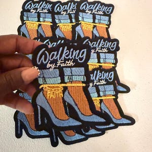 May include: An embroidered patch featuring a pair of light blue high-heeled shoes with cuffed blue jeans, a gold ankle chain with the word FAITH, and the text Walking by Faith in blue script lettering against a black background.