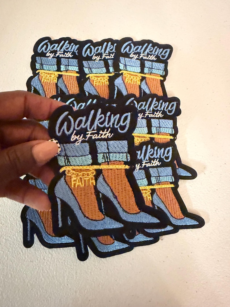 May include: An embroidered patch featuring a pair of light blue high-heeled shoes with cuffed blue jeans, a gold ankle chain with the word FAITH, and the text Walking by Faith in blue script lettering against a black background.