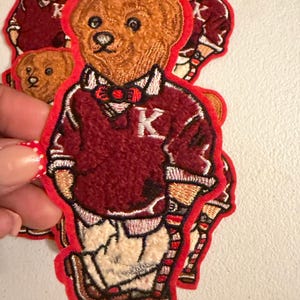 May include: An embroidered patch featuring a brown teddy bear wearing a maroon sweater with a white letter K, a white collared shirt, a red bow tie, and cream-colored trousers. The patch has a bright red border and a textured, stitched finish.