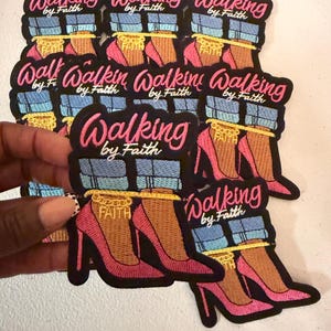 May include: A collection of embroidered patches featuring a graphic of legs in blue jeans and pink high heels with a gold ankle chain that says FAITH. The text Walking by Faith is embroidered in pink and white above the legs on a black background.