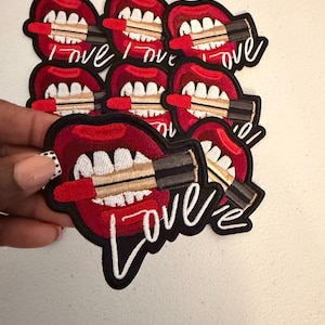 May include: A collection of embroidered patches featuring a pair of red lips with white teeth holding a red lipstick tube. Each patch has a black border and the word Love embroidered in white cursive script at the bottom of the design.