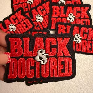 May include: A collection of embroidered patches featuring the text "BLACK & DOCTORED" in bold red capital letters with a white ampersand symbol. Each patch has a thick black border and a textured fabric finish, showcasing the same design.