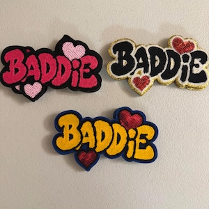 May include: Three embroidered patches featuring the word BADDIE in a bold, stylized font. The top left patch is pink with light pink hearts, the top right is black with red hearts and gold trim, and the bottom patch is yellow with red hearts and blue trim.