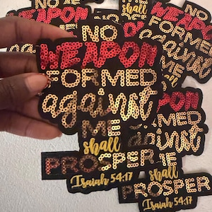 May include: Black patches with gold and red sequin lettering. The patches feature the text "NO WEAPON FORMED AGAINST ME SHALL PROSPER" and "Isaiah 54:17". The patches are held in a hand.