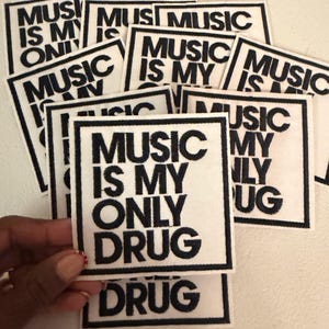 May include: A collection of white square patches with black text that reads "MUSIC IS MY ONLY DRUG". The patches are surrounded by a black border. The patches are stacked on top of each other.