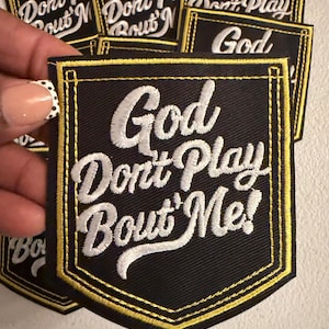 May include: A black fabric patch shaped like a pocket with yellow embroidered borders. The text God Don't Play Bout' Me! is embroidered in white script across the center. The patch features a pointed bottom edge and a rectangular top section.