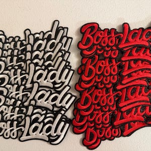 May include: A collection of embroidered patches featuring the text Boss Lady in a cursive script font. The patches are arranged in two distinct groups, with one set featuring white lettering on a black background and the other set featuring red lettering on a black background.