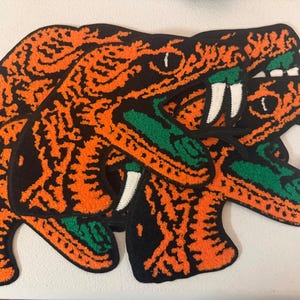 May include: Three snake-shaped rugs with open mouths, featuring orange and black patterns, green tongues, and white fangs. The rugs are made of a textured material, possibly fabric or yarn, and are arranged in a layered composition.