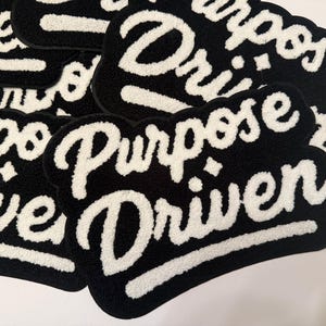 May include: A collection of black and white patches with the words "Purpose Driven" in a cursive font. The patches have a soft, textured appearance, with the white text standing out against the black background. The patches are arranged in a slightly overlapping pattern.