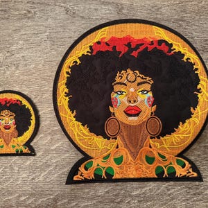 May include: Two embroidered patches featuring a Black woman with a large afro. The larger patch is circular with a gold border and a detailed face with colorful accents. The smaller patch mirrors the design. The woman has large hoop earrings and a red lip.