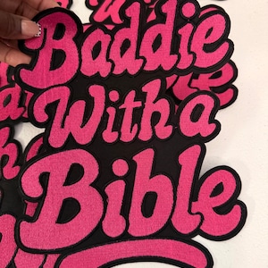 May include: A black fabric patch featuring the embroidered text Baddie With a Bible in bright pink thread. The lettering is in a bold, cursive script style with a thick black outline, creating a layered appearance for the embroidered design.