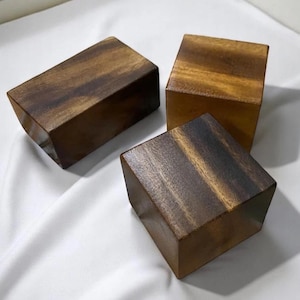 Puede incluir: Tres bloques de madera con un rico patrón de vetas marrón oscuro. Dos son rectangulares y uno es cuadrado. Los bloques están dispuestos sobre una superficie blanca, con sombras que añaden profundidad. La madera tiene un acabado pulido, resaltando las variaciones naturales de color.